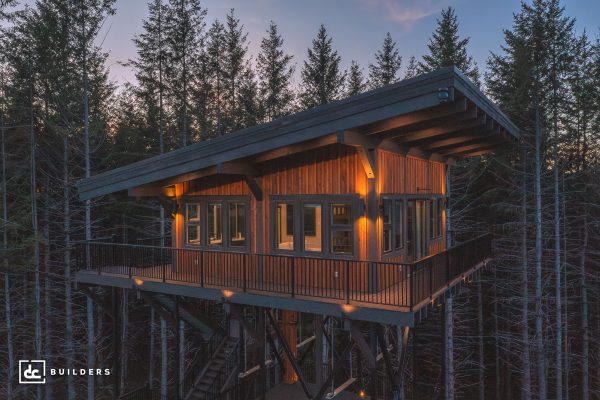 A treehouse-style building with a wide balcony and large windows, elevated on metal supports, surrounded by trees at dusk.